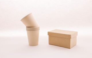 Kraft paperboard box with lid beside a stack of paper cups on a light background